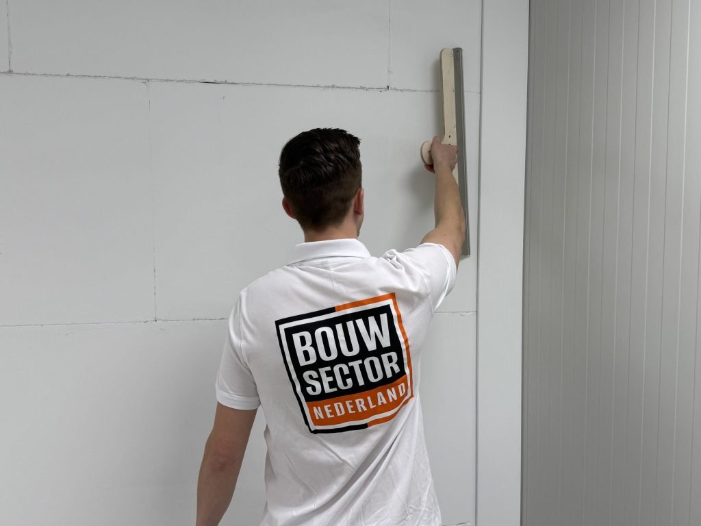 Stukadoor van Bouwsector Nederland in wit poloshirt met logo houdt een lange spaan tegen de muur