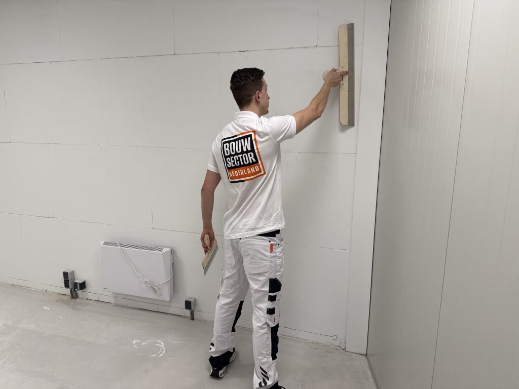 Stukadoor van Bouwsector Nederland trekt een wand vlak met een lange spaan, een plakspaan in zijn andere hand