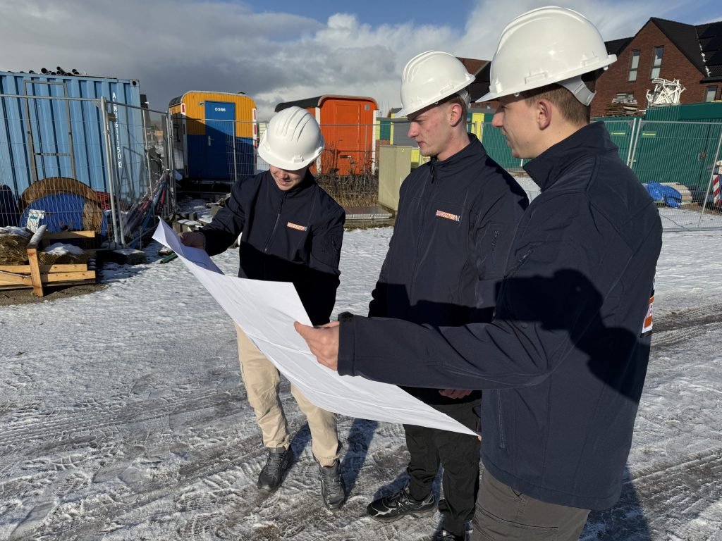 Drie vakmensen van Bouwsector Nederland met witte helm bekijken een bouwtekening op een nieuwbouwterrein